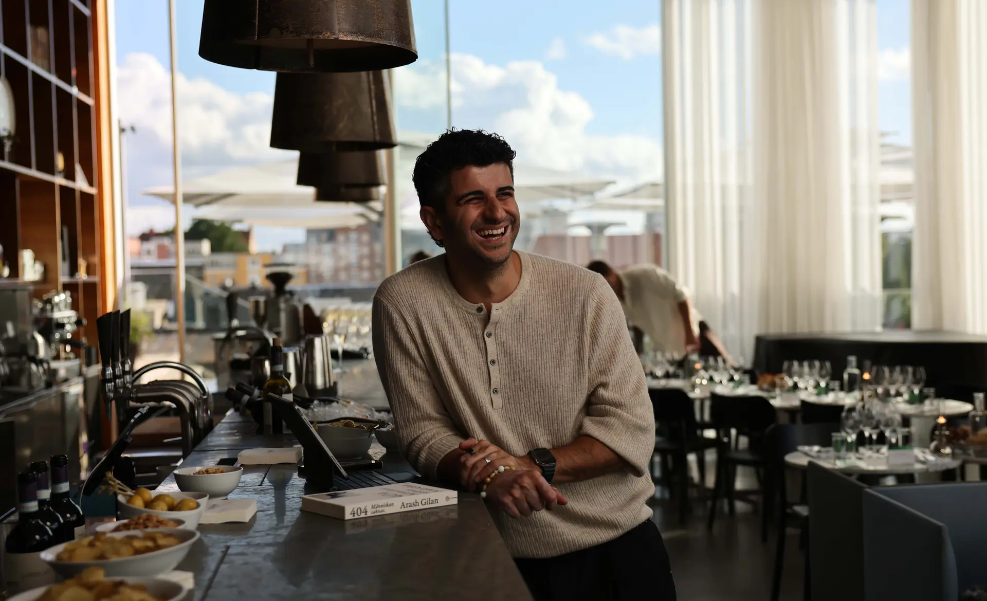 A man in a beige sweater smiles while leaning on a bar counter in a bright, modern restaurant with city views.