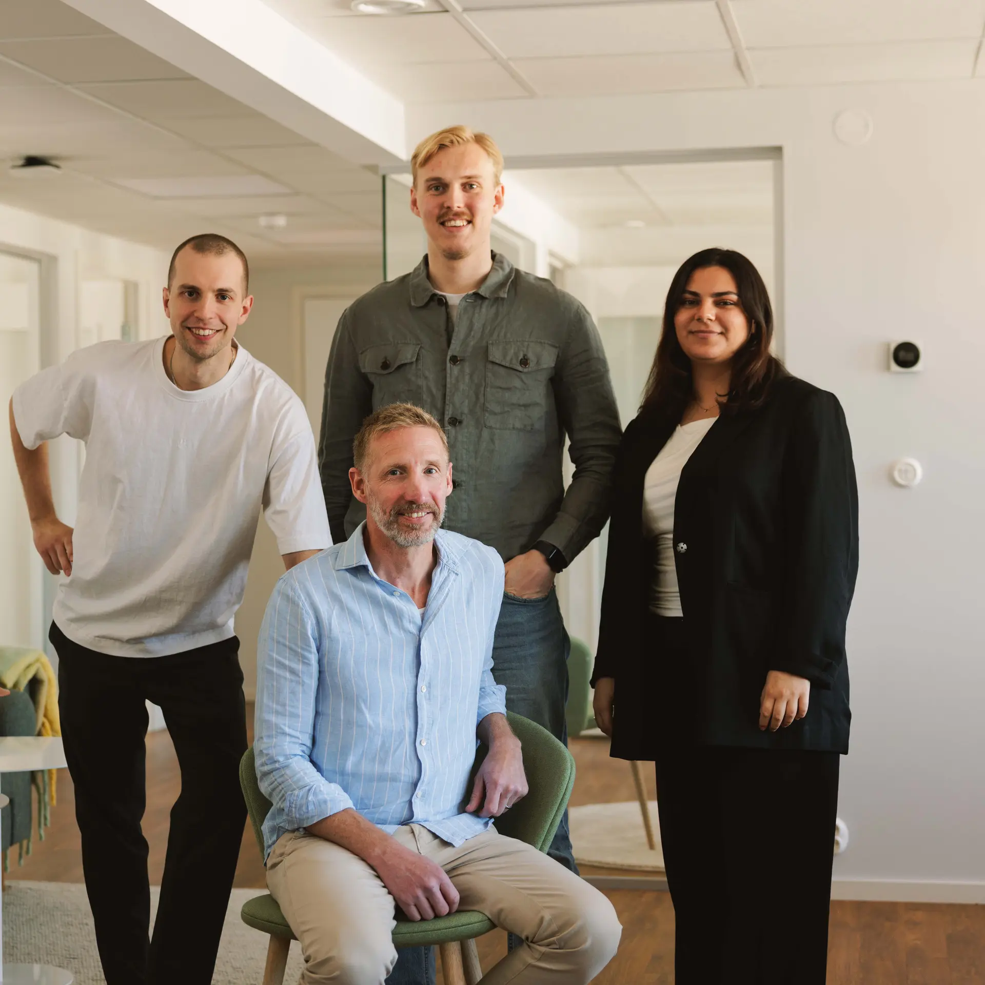 Four people posing in a modern office; one seated, three standing. They appear friendly and professional, with casual and business attire.