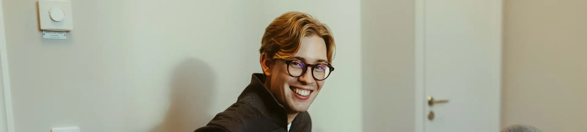 A smiling person with glasses and light brown hair, wearing a dark jacket, sits in a room with a thermostat on the wall.