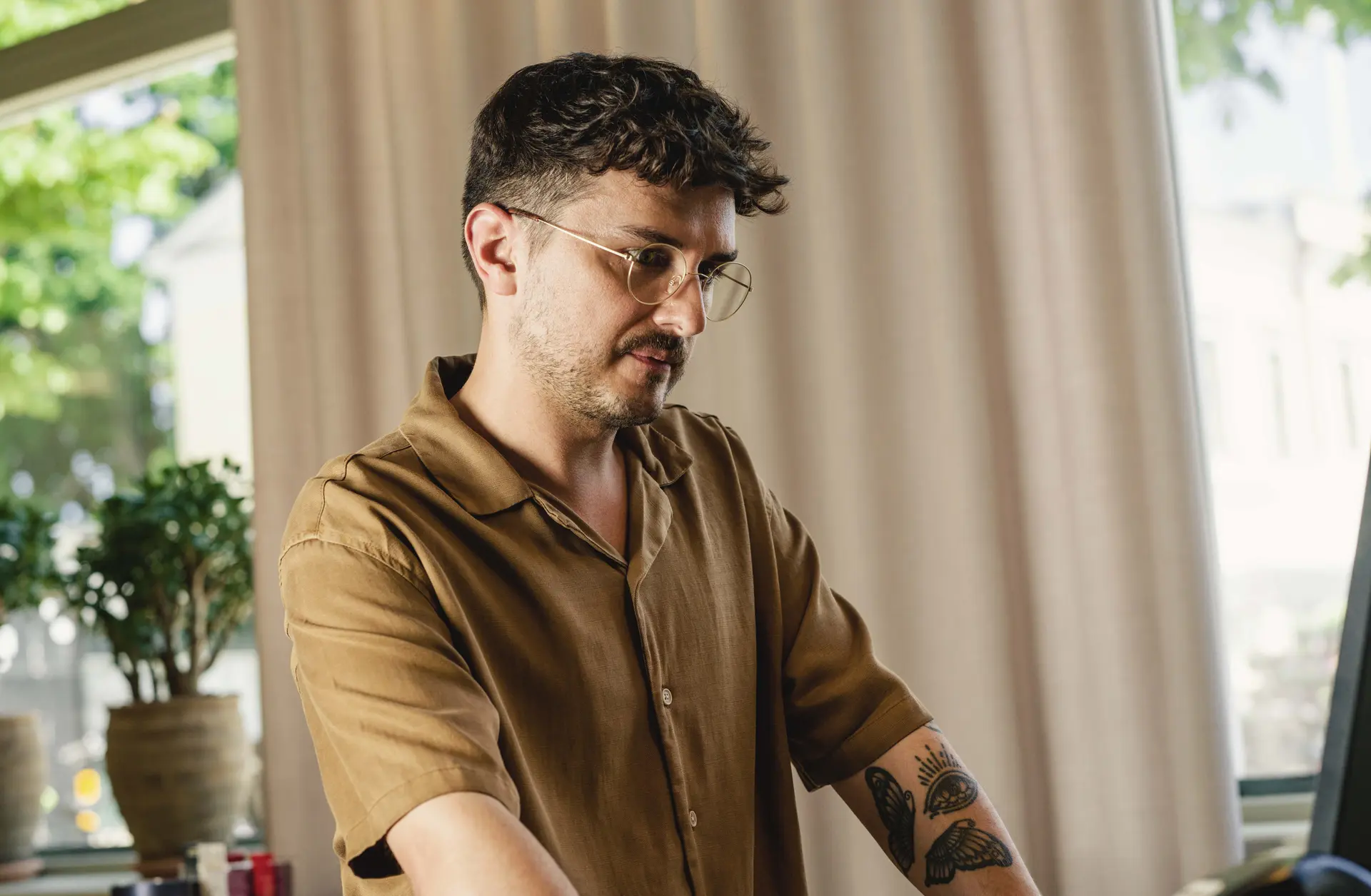 A man with glasses and a brown shirt works on a computer, with a tattoo visible on his arm and a potted plant in the background.
