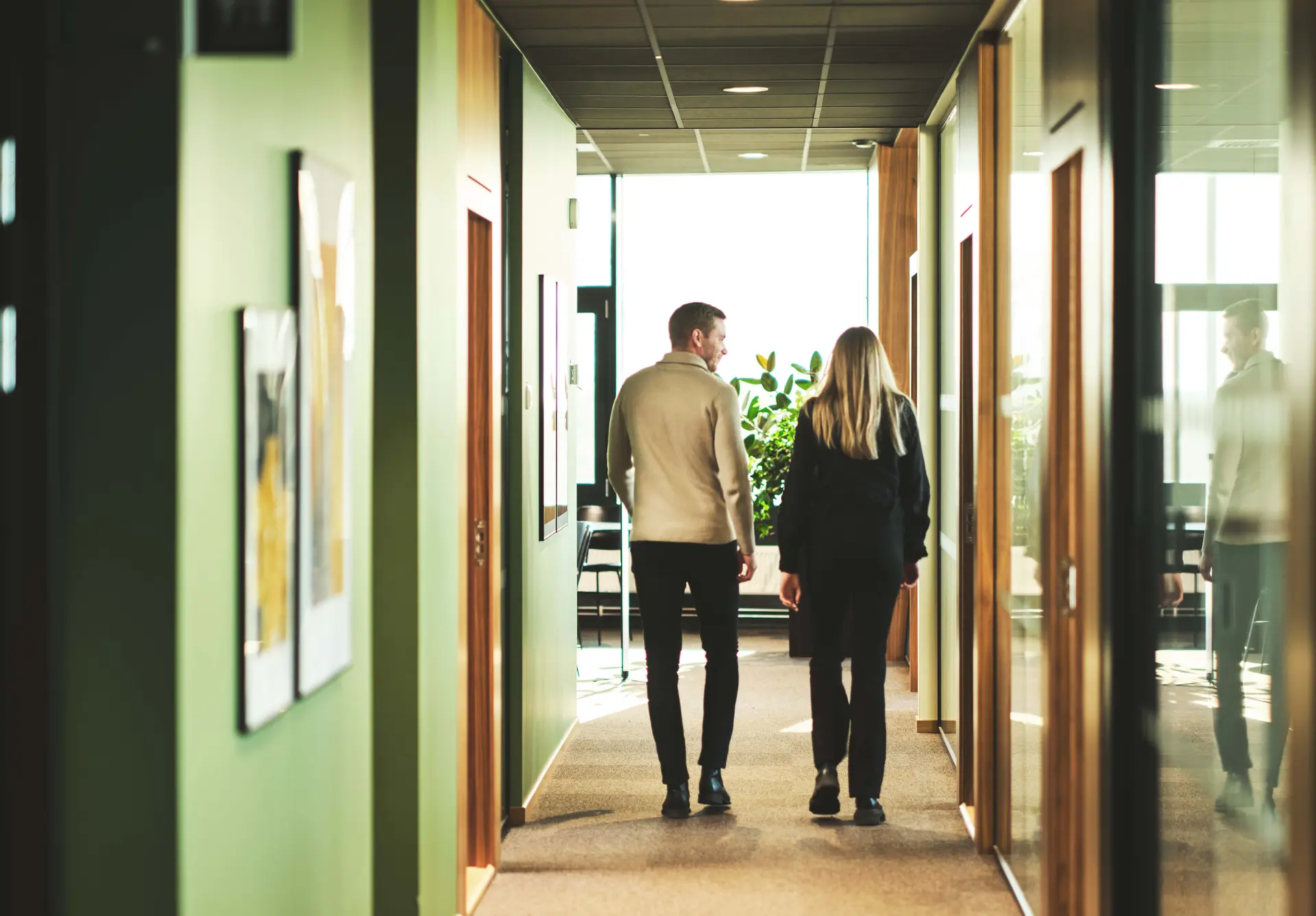Two people walk down a sunlit corridor with green walls and large windows, with plants visible at the end.