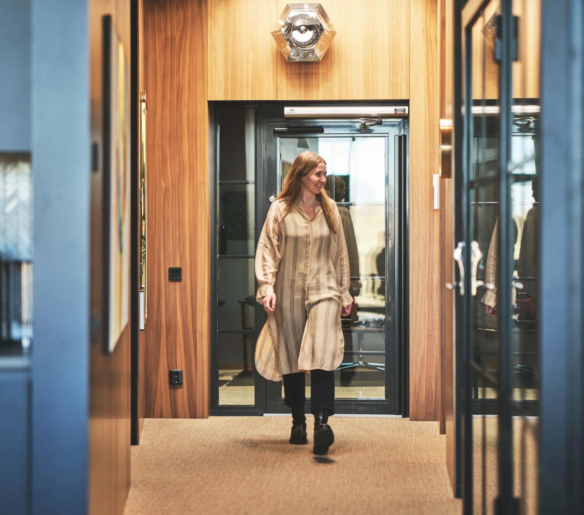A woman in a long beige dress walks through a modern office hallway with wood paneling and glass doors.