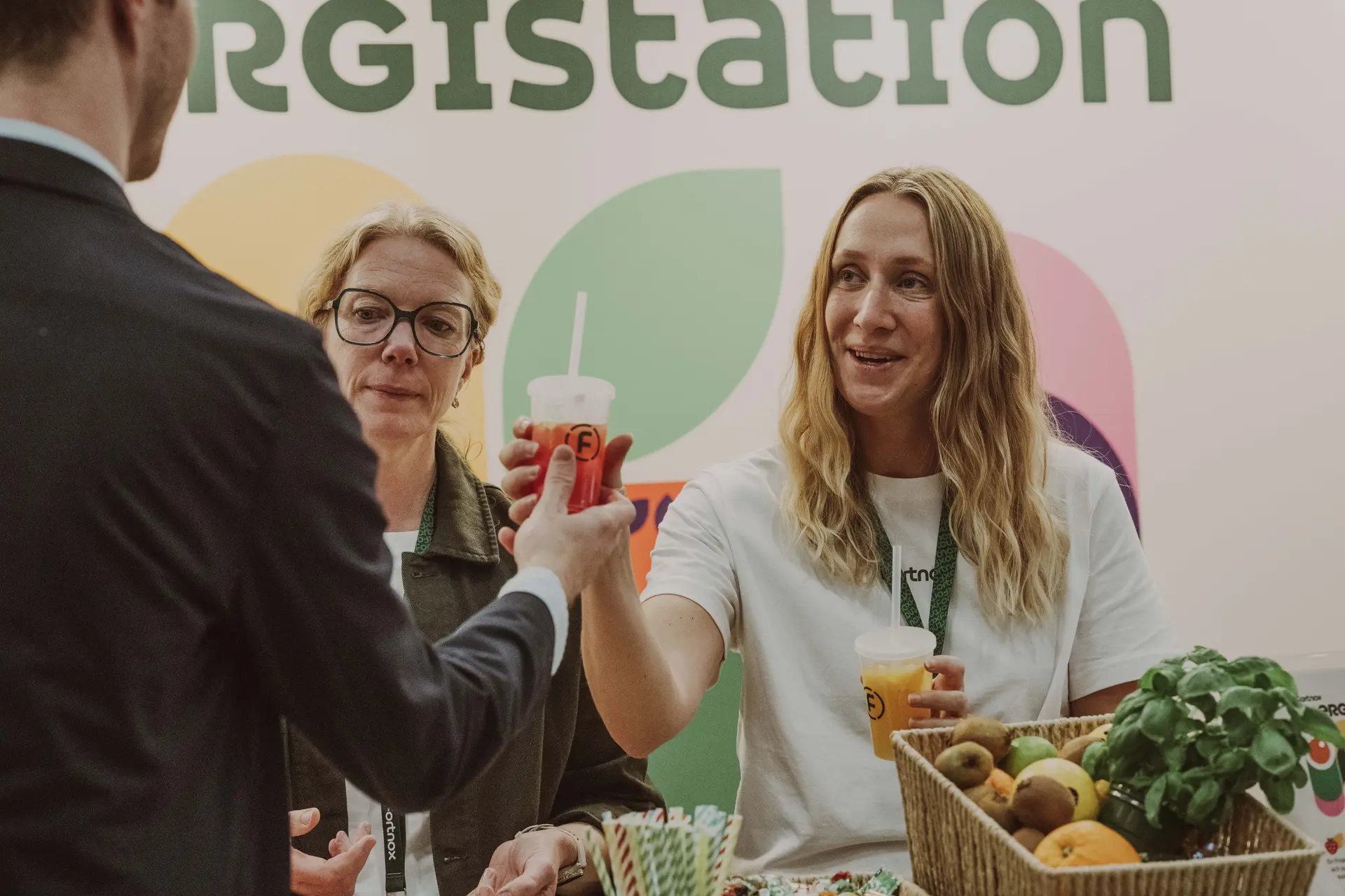 Two women at a booth handing drinks to a man. A basket of fruit and a plant are on the table. Large colorful signage is in the background.