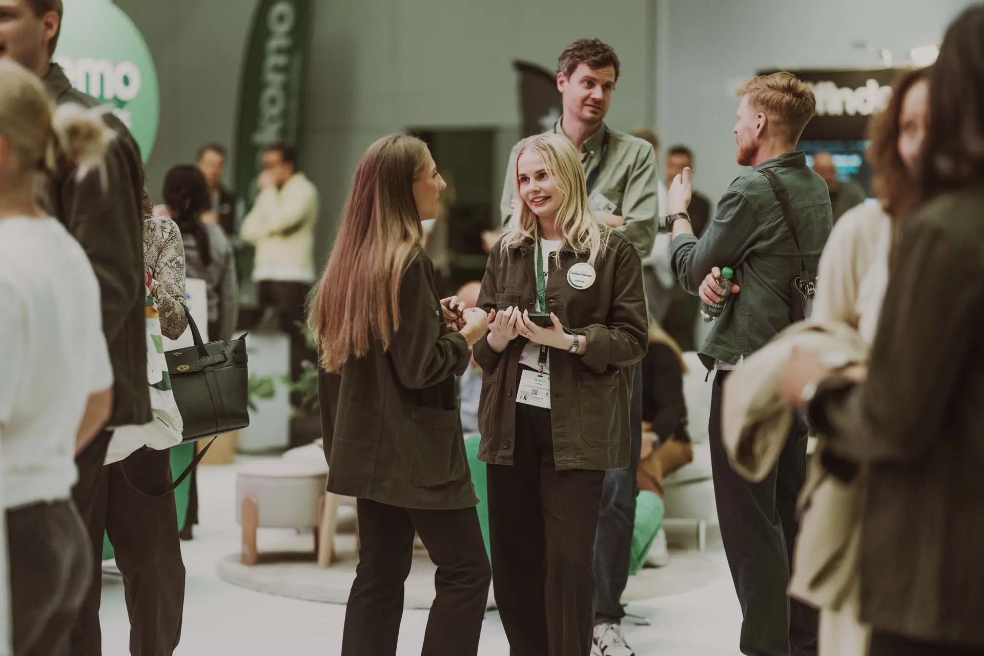 People mingling and conversing at a networking event, with a focus on a woman holding a phone and badge, surrounded by others in casual attire.
