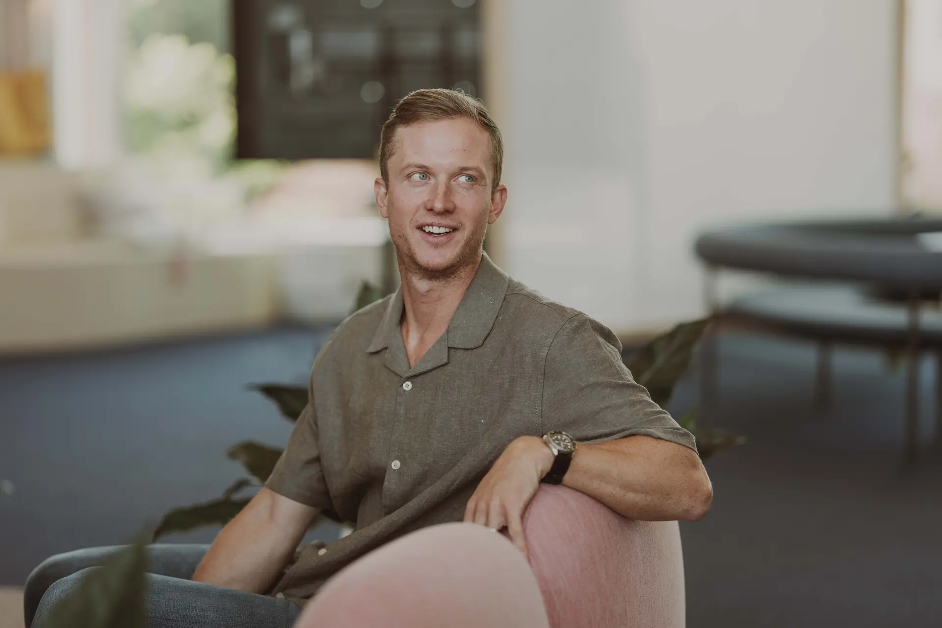A man in a casual shirt sits on a pink chair, smiling and looking to the side, in a bright room with blurred background.