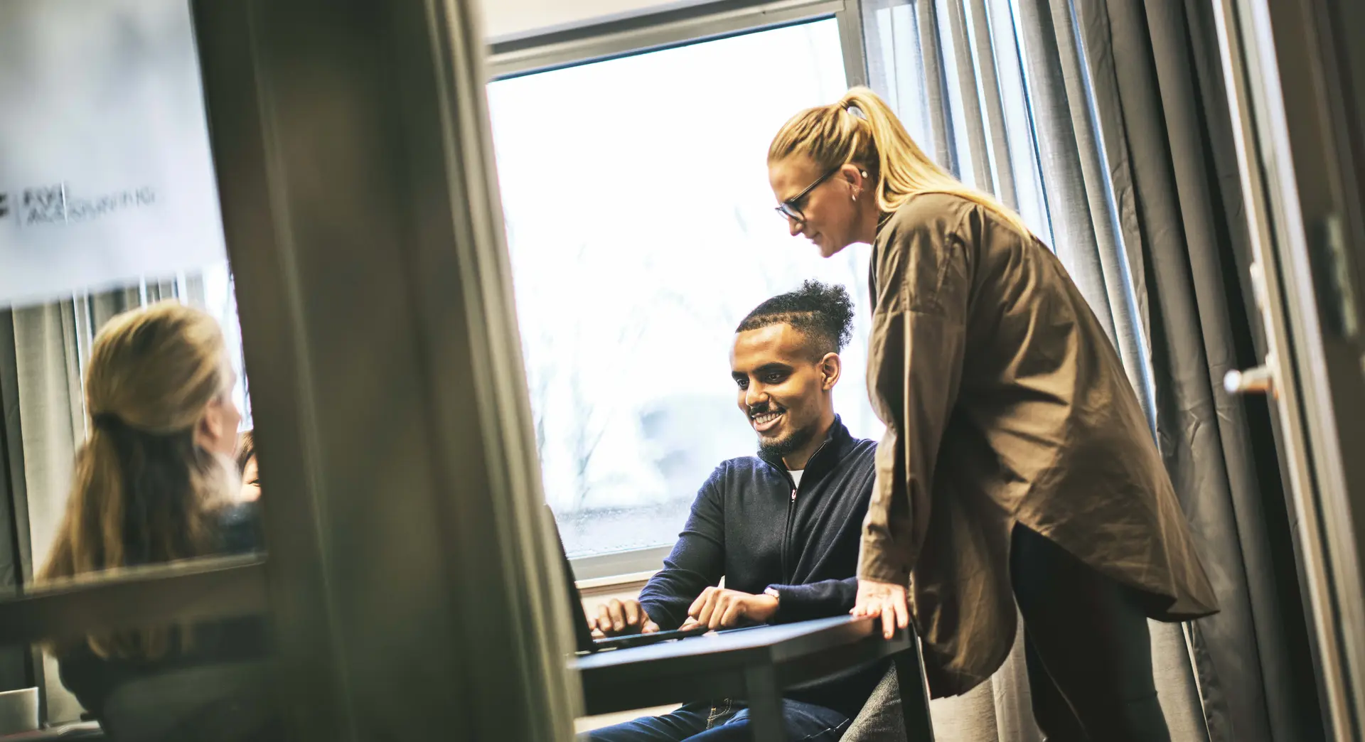 Two people collaborating in an office by a window, with one person seated smiling and typing on a laptop while the other stands and observes.