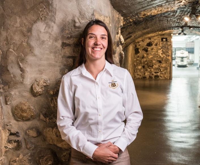 A woman in a white shirt, smiling, stands in a stone-walled cellar or tunnel with warm lighting.