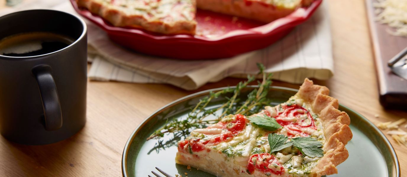 A slice of vegetable quiche with tomatoes and herbs on a green plate, garnished with fresh thyme, beside a black mug and a whole quiche.