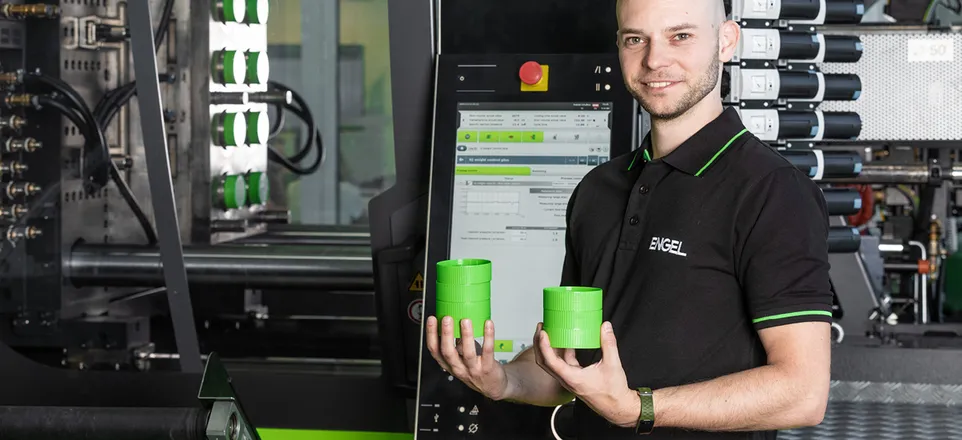Image shows development engineer Matthias Hüttler in front of an ENGEL injection moulding machine with two green injection moulded components in his hand