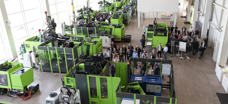 Image shows a view into the ENGEL technical center with numerous green ENGEL injection molding machines and an interested audience