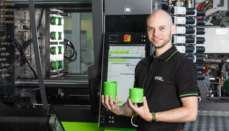 Bild zeigt Entwicklungsingenieur Matthias Hüttler vor einer ENGEL Spritzgießmaschine mit zwei grünen Spritzgussbauteilen in der Hand