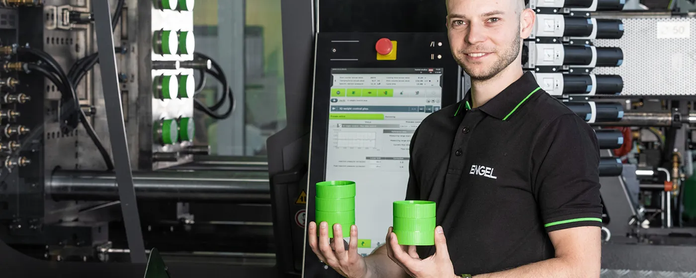 Image shows development engineer Matthias Hüttler in front of an ENGEL injection moulding machine with two green injection moulded components in his hand