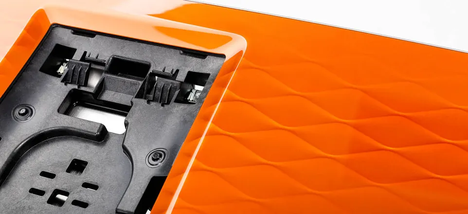 Nahaufnahme eines schwarzen Kunststoffbauteils mit orangefarbener, im clearmelt-In-Mould-Coating-Verfahren beschichteter Oberfläche.