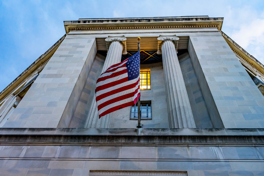 Low-angle view of a neoclassical building with tall columns, featuring an American flag waving in the foreground under a cloudy sky.