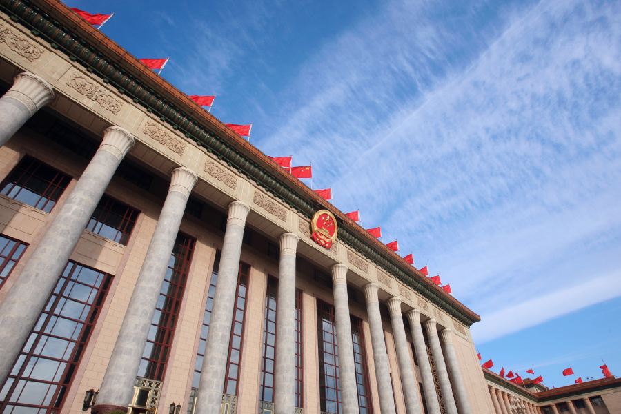 Exterior of the Hall of the People building in Beijing, featuring tall columns, red flags on the roof, and clear blue sky.