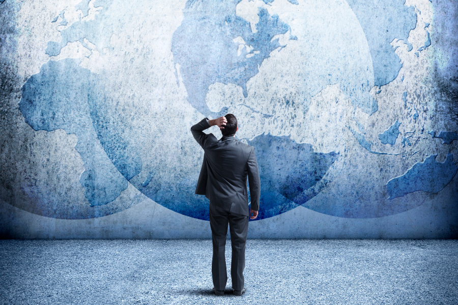 Man in a suit stands on a textured floor, looking at a large blue and white world map on a wall, appearing thoughtful and puzzled.