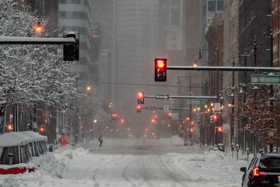 Snow-covered city street with red traffic lights glowing, blanketed cars, and a person walking in the distance on a foggy winter day.