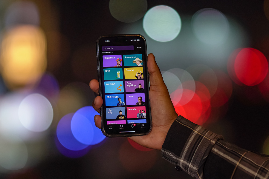 A hand holding a smartphone displaying a colorful music streaming app interface, with a blurred city nightscape in the background.