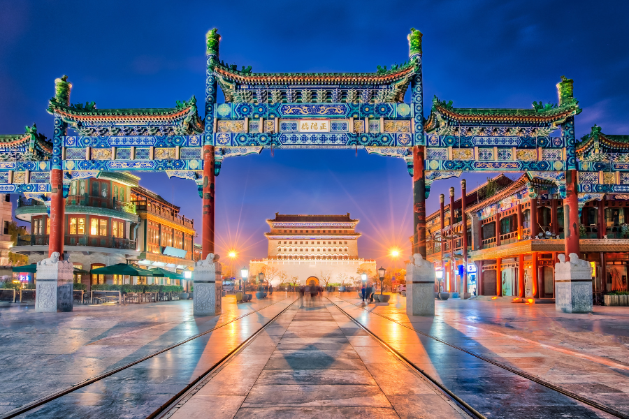 Illuminated traditional Chinese gateway and buildings at night in Beijing, with a lit-up tower visible through the archway.