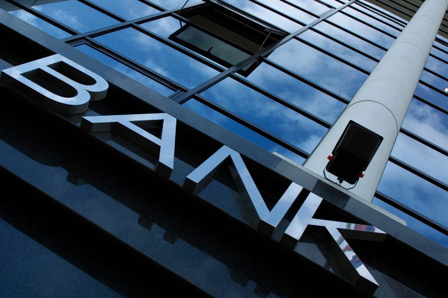 Close up photo angled up towards a "BANK" sign on the side of a tall glass building, reflecting blue sky and clouds
