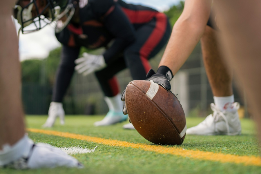 A close-up of a football on the ground ready for a snap, with players in the background poised to start the play on a green field.