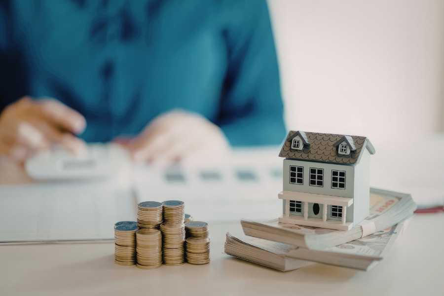 Stacks of coins and a small house model on documents, with a person in blue using a calculator in the background.