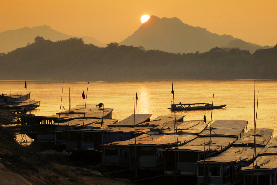 Boats docked along the Mekong river at sunset, with silhouetted mountains in the background and a golden sky.