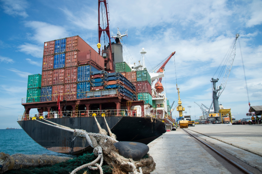 A large cargo ship loaded with colorful shipping containers docked at a busy port, surrounded by cranes and workers under a partly cloudy sky.