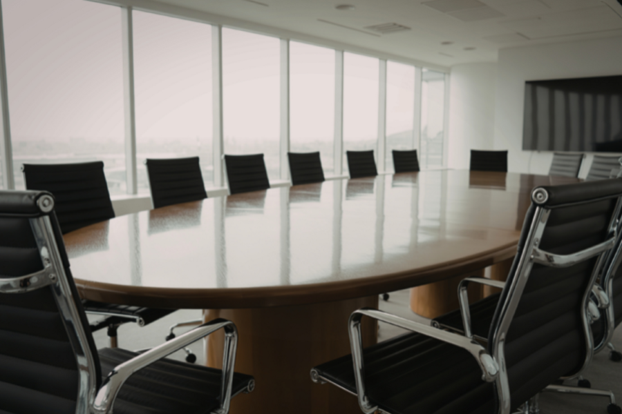 A modern conference room with a large oval table surrounded by black leather chairs, facing large windows and a wall-mounted TV.