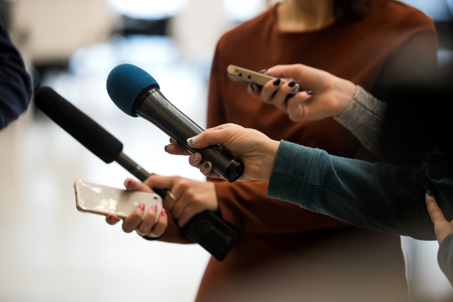 Several hands holding microphones and a smartphone, directed toward an unseen person during an interview.
