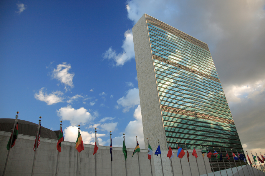 The UN Headquarters in NYC, windows reflecting clouds, surrounded by flags from various countries on poles, under a partly cloudy sky.