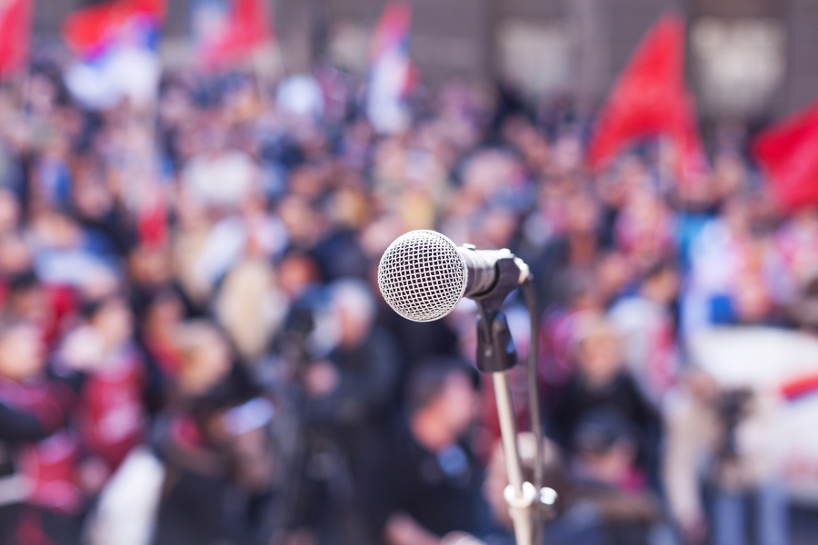 Close-up of a microphone in focus with a blurred crowd and red flags in the background, suggesting a public political speech