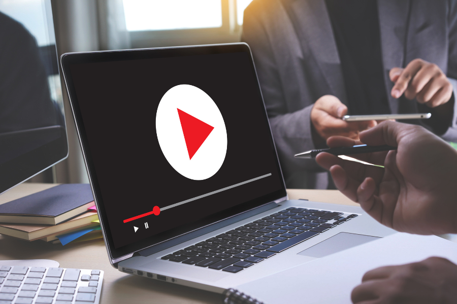 Laptop screen showing a paused video with a large play button. Hands in the background hold a pen and smartphone.