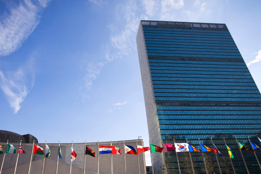 A tall modern glass building with numerous international flags flying in front, set against a clear blue sky.
