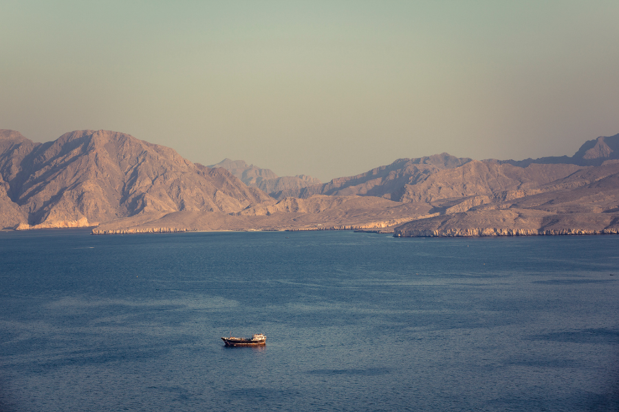 A small boat floats on a calm blue sea in the Strait of Hormuz with rugged, sunlit mountains in the background under a clear sky.