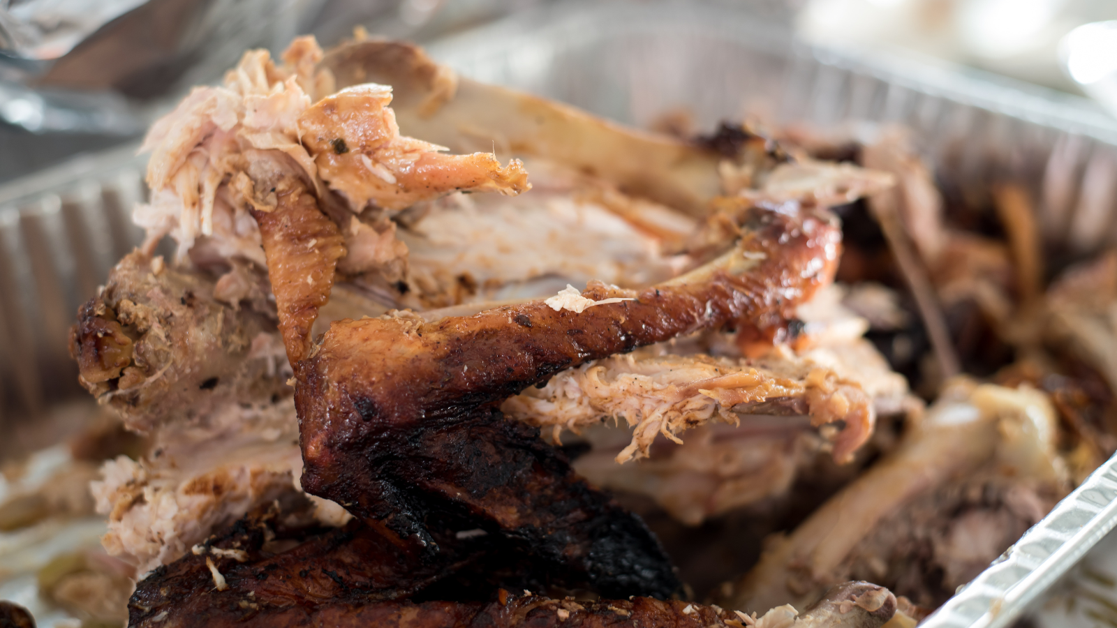 Pile of carved turkey bones and shredded browned meat with charred skin in an aluminum serving tray.