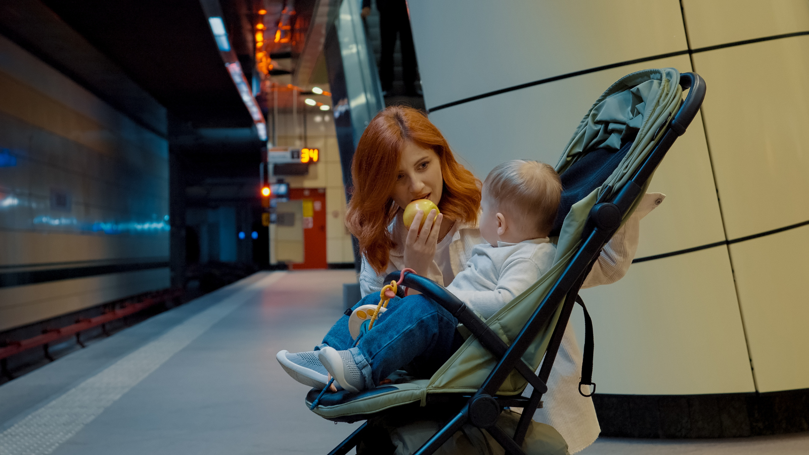 Woman with red hair in a subway station, feeding an apple to a baby in a stroller, with a train platform in the background.