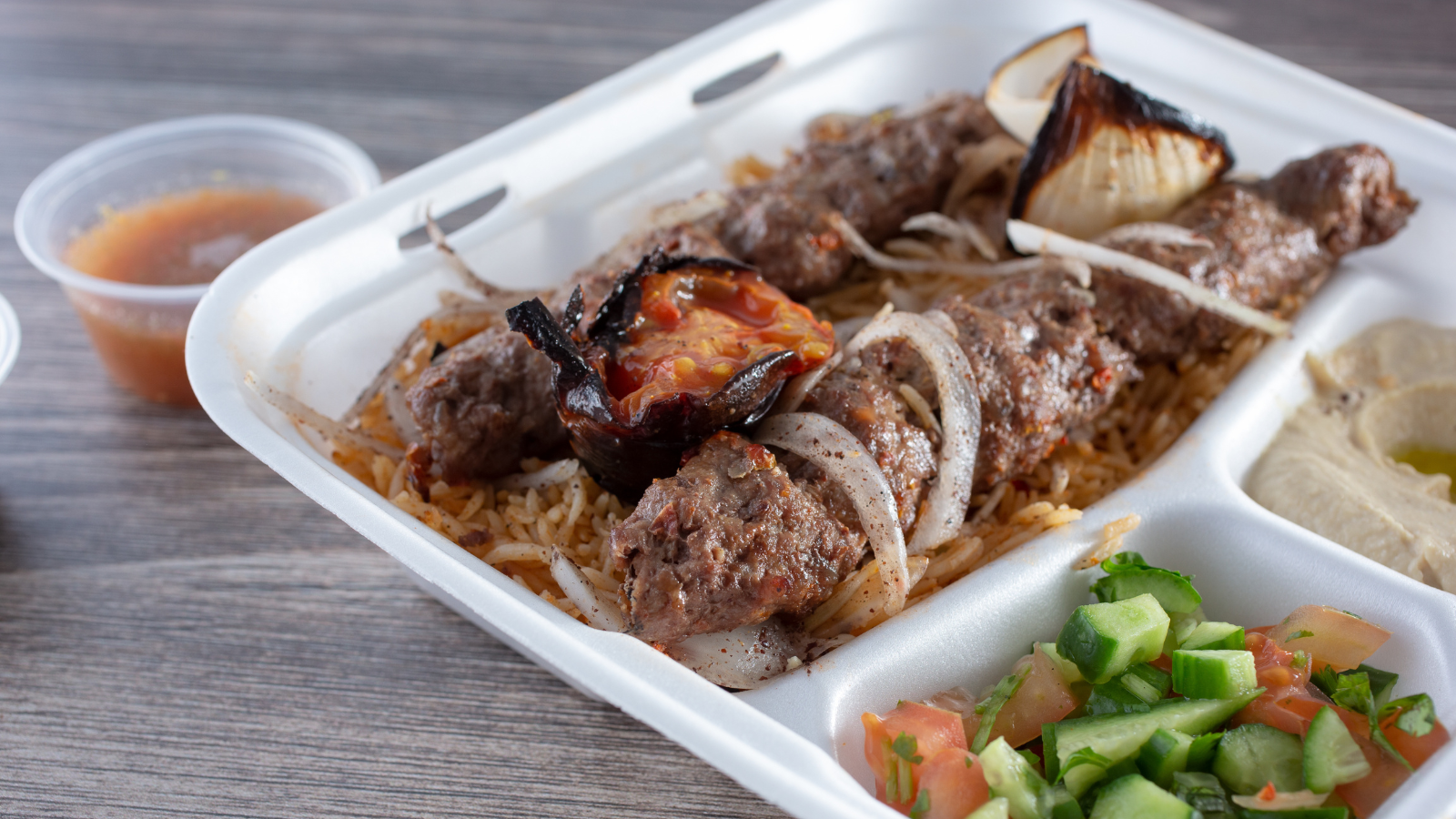 A takeout container with grilled kebabs on rice, roasted onion, hummus, and a cucumber-tomato salad, accompanied by a small sauce cup.