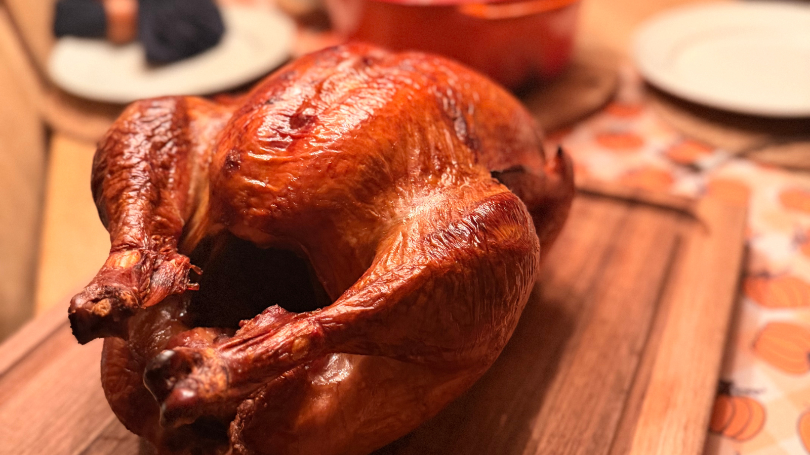 Golden-brown roasted whole turkey on a wooden cutting board, set on a table with plates and pumpkin-patterned tablecloth.