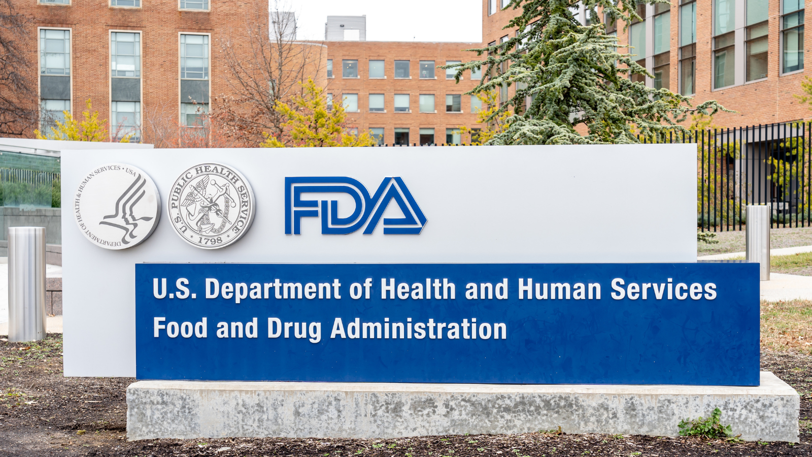 Exterior view of the U.S. Food and Drug Administration (FDA) headquarters featuring a prominent sign with the FDA logo and the text "U.S. Department of Health and Human Services Food and Drug Administration" in front of a brick office building.