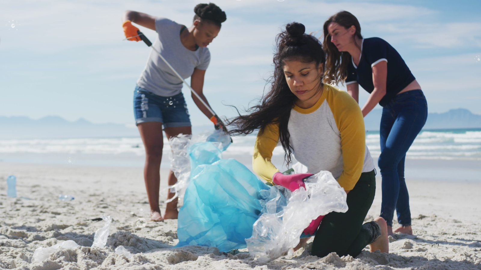 Three female volunteers clean up plastic waste on a sandy beach. They are focused and wearing casual clothes with gloves. The ocean is visible in the background.