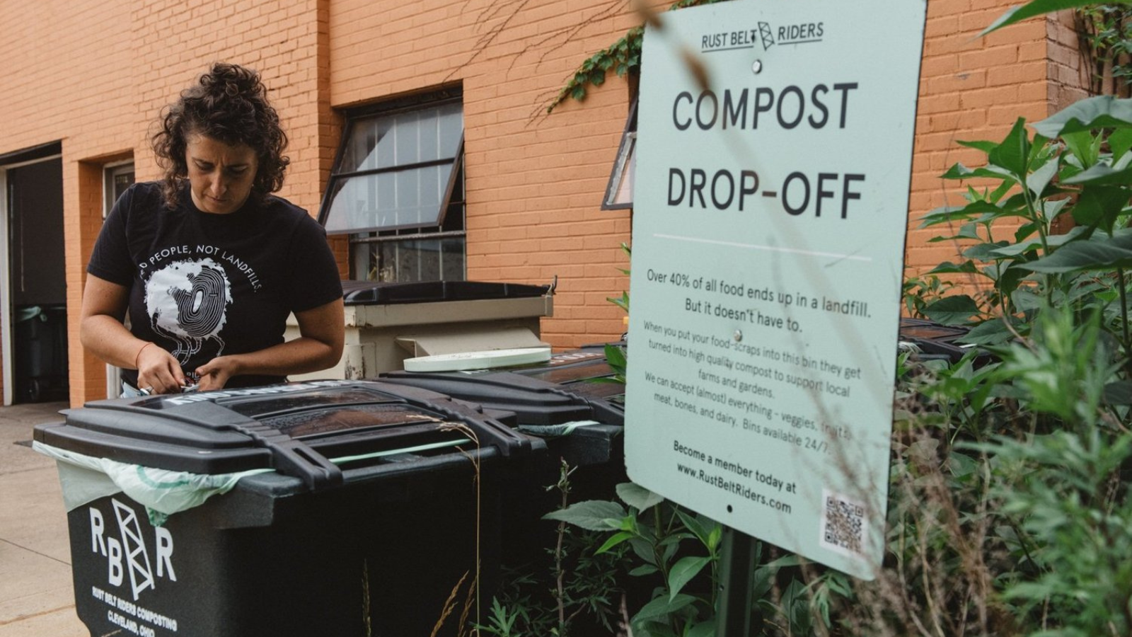 A Rust Belt Riders worker facilitates compost drop-off at a designated collection site in Cleveland, Ohio. The worker, wearing a "Feed People, Not Landfills" t-shirt, secures a compost bin next to a sign explaining the importance of food waste diversion. The image highlights community-driven composting efforts and the role of Rust Belt Riders in promoting sustainability.