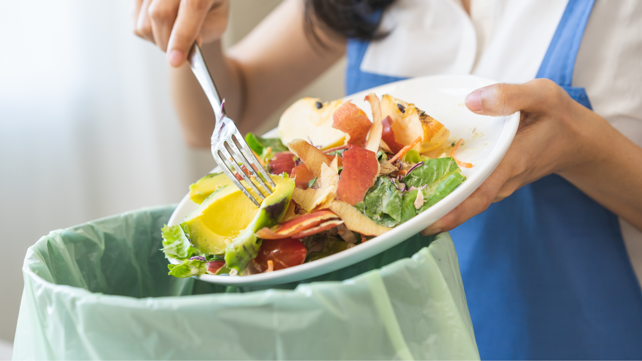 Person scraping food scraps from a plate into a trash bin with a green compost bin-liner, wearing a blue apron.