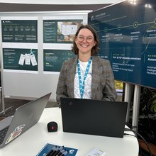 A enox-share person smiling at a tech booth with laptops, promotional brochures, and a display board featuring diagrams and information.