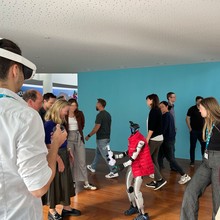 A group of people, some wearing lanyards, observe a small humanoid robot in a red vest inside a room with a blue wall.