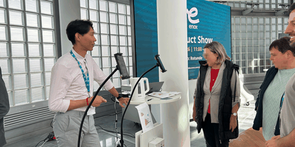A person demonstrating a product at a trade show booth to a group of attentive attendees in a bright, modern venue.