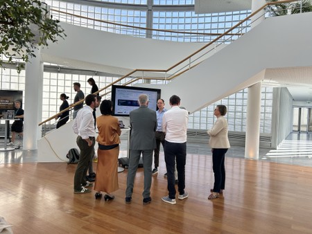 A group of people discuss while standing near an enox presentation screen in a modern, sunlit atrium with a curved staircase and glass block wall.