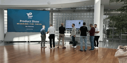 Seven people stand in a modern space with wooden floors, a large screen displaying an announcement for a product show on 28.09.2026.