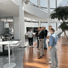 A group of people stands in a modern, bright office space discussing in front of a large monitor. The room features a tree and curved glass walls.