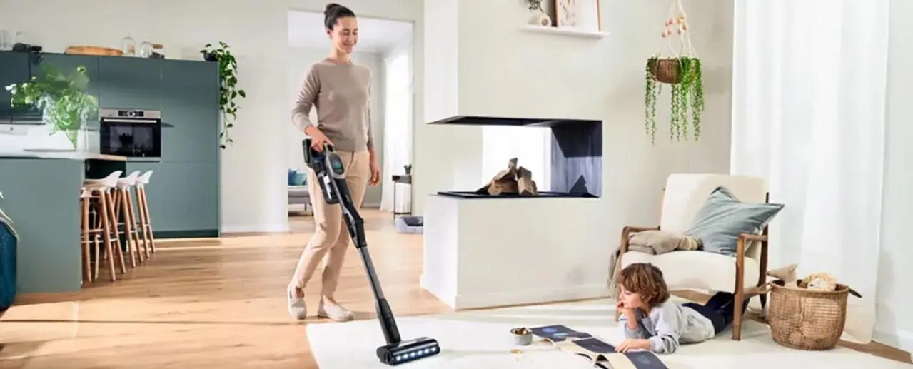 Jonge vrouw stofzuigt de huiskamer met een Bosch steelstofzuiger. Kind ligt op de grond een boek te lezen.